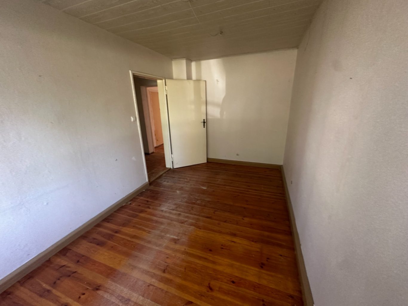 Leerer Raum mit Holzboden und offener Tür, weiße Wände.