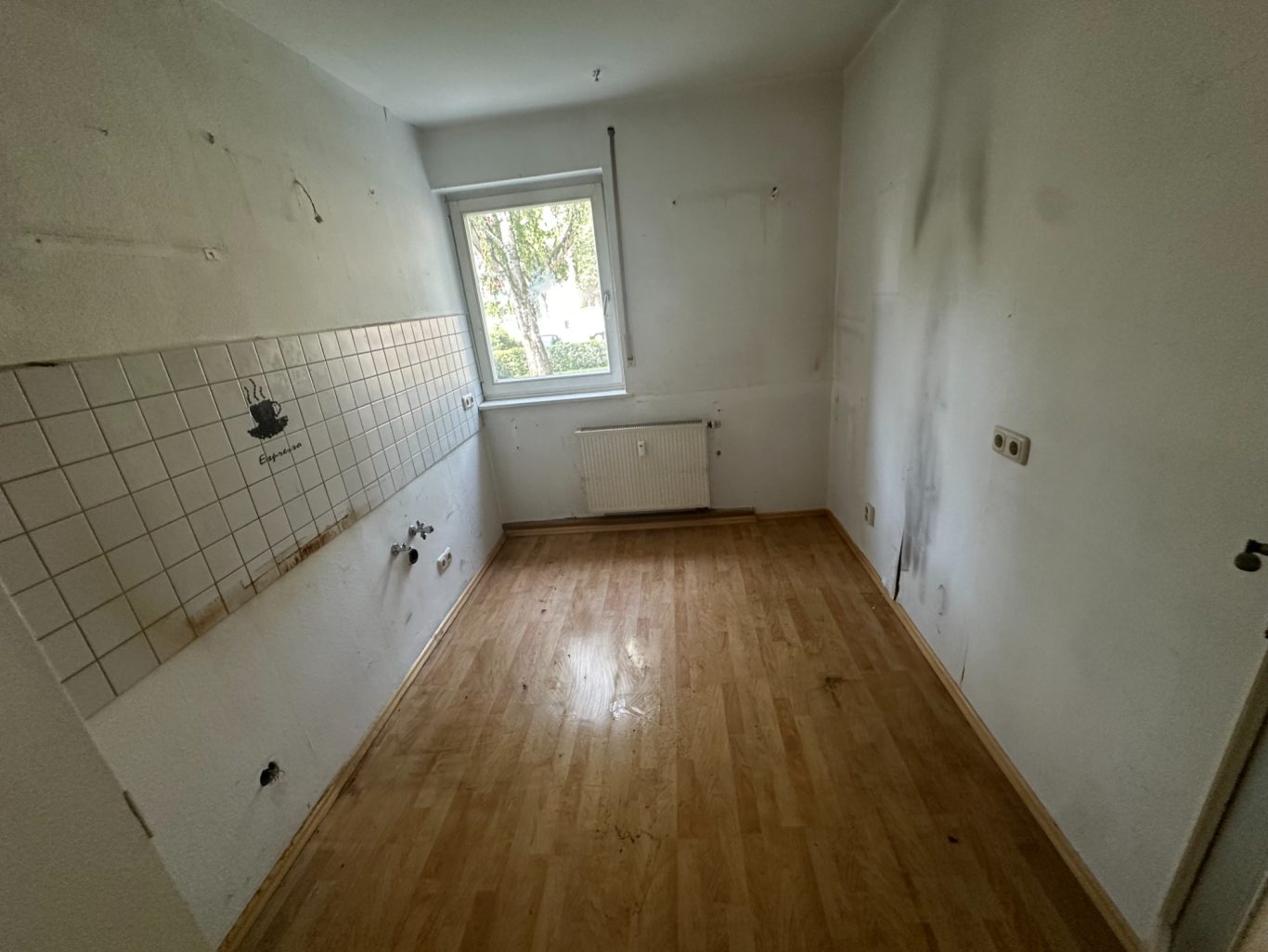 Leere, unrenovierte Küche mit Holzboden und Fenster, teilweise beschädigte Wände.